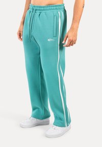 Pantalons de survêtement turquoise avec taille élastique à cordon, poches latérales, bandes blanches sur les côtés et un petit logo près de la poche. Texture lisse.