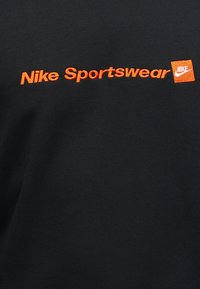 Λογότυπο κειμένου Nike Sportswear σε έντονο πορτοκαλί με ένα μικρό πορτοκαλί τετράγωνο με το λευκό λογότυπο Nike σε μαύρο ύφασμα.