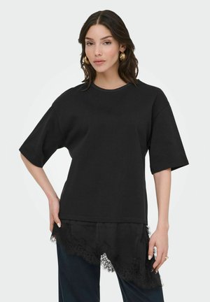 Donna con capelli mossi e scuri indossa una maglia nera a maniche corte, ampia, con orlo in pizzo asimmetrico e pantaloni scuri, posando con una mano sul fianco.