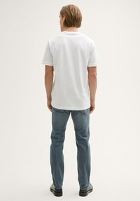 Fehér, rövid ujjú, kerek nyakú pamut póló világoskék farmernadrággal és fekete cipővel, hátulról nézve.