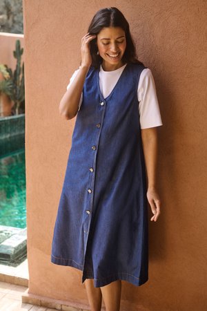 Femme souriante touchant ses cheveux, portant une robe en jean sans manches boutonnée sur une chemise blanche à manches courtes, debout près d'un mur en terre cuite.