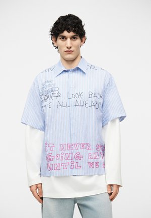 Jeune homme aux cheveux bouclés et foncés portant une chemise à manches courtes rayée bleue par-dessus un haut à manches longues blanc, avec un texte écrit à la main sur la chemise.