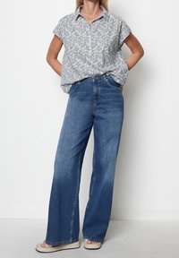 Puuvillapaita, jossa harmaa kukkakuosi, lyhyet hihat ja rento istuvuus, yhdistetty leveälahkeisiin sinisiin farkkuihin ja vaaleisiin sandaaleihin.