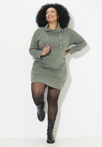 Vestido sudadera verde oliva con escote de capucha y cordón, mangas largas y bolsillos laterales, combinado con botas de tobillo negras y medias transparentes.