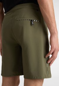 Shorts verts en tissu lisse, avec un motif de rayure latérale et une poche arrière dotée d'une languette de tirage à carreaux noirs et blancs.