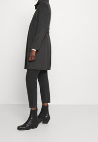 Ciemnoszary wełniany płaszcz z wysokim kołnierzem, zwężoną talią, bocznymi kieszeniami, połączony z czarnymi dżinsami i błyszczącymi czarnymi botkami na kostkę.