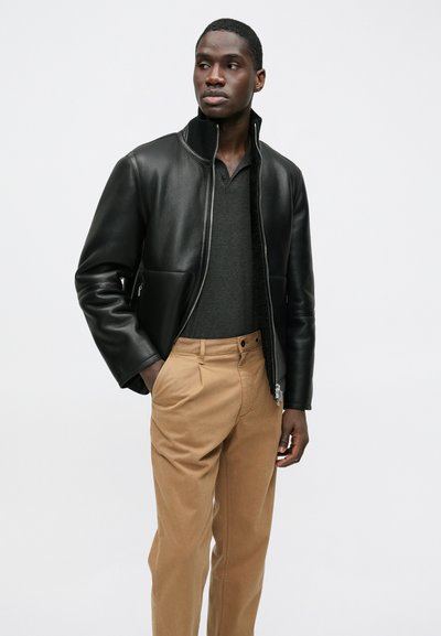 Black leather bomber jacket with a high collar, two front pockets, paired with beige trousers and a dark gray polo shirt.