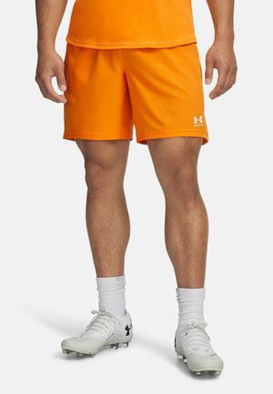 Hombre vestido con pantalones cortos deportivos de color naranja brillante, calcetines blancos y botas blancas con logotipos negros de Under Armour, de pie sobre un fondo blanco.
