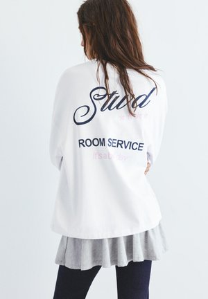 Femme aux longs cheveux bruns, portant une veste blanche avec les inscriptions « Stud », « ROOM SERVICE » et « It's a holiday » au dos, par-dessus une jupe grise.