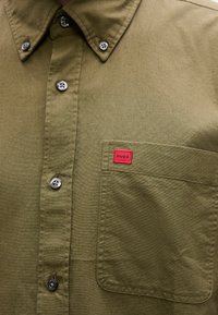 Πουκάμισο σε ελιά πράσινο, φτιαγμένο από ύφασμα, με τσέπη στο στήθος που φέρει κόκκινη  ετικέτα "HUGO" και ασημένια κουμπιά. Λείο υφή.