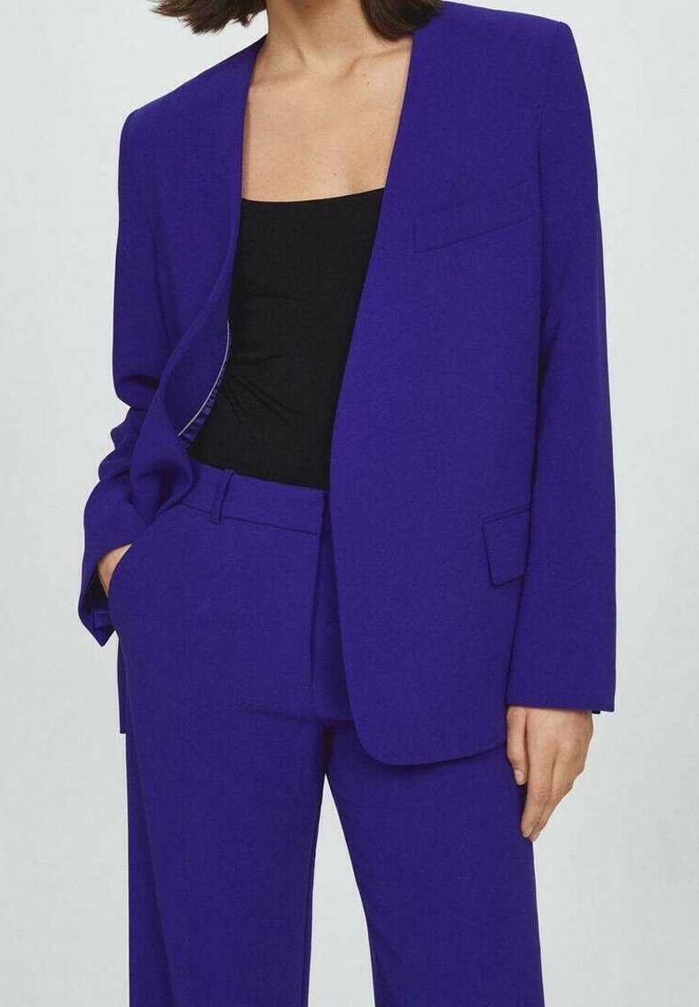 Femme portant un tailleur-pantalon violet vif avec un haut noir, posant avec une main dans la poche sur un fond clair uni.
