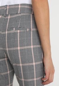 Close-up van een persoon die hoge taillebroeken draagt met een zwart-wit-zachte roze ruitpatroon en een achterzak, hand ontspannen naast zich.