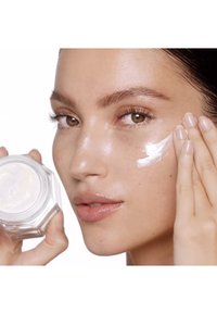 Clear glass cream jar held in one hand, with a model applying a smooth, white cream on her cheek. Skin appears healthy and hydrated.