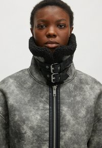 Grau gefütterte Kunstpelzjacke mit hohem Kragen, schwarzen Akzenten, doppelten Schnallen und einem Frontreißverschluss. Strukturierte Oberfläche mit einem gesprenkelten Muster.