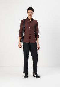 Cămașă maro cu nasturi, cu mâneci lungi, asociată cu pantaloni negri și adidași negri cu dungi albe. Geantă neagră purtată pe umăr.