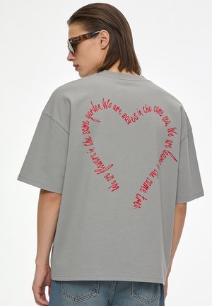 Persona che indossa una maglietta grigia oversize con testo a forma di cuore rosso sulla schiena e occhiali da sole, di profilo.
