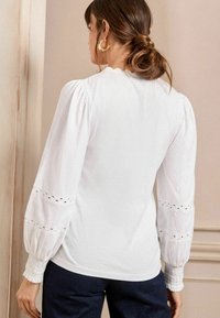 Blusa bianca con maniche a sbuffo lunghe, caratterizzata da dettagli in pizzo ai polsini e un orlo arricciato. Tessuto liscio e design aderente in vita.