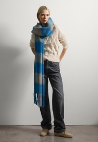 Bufanda de lana con un patrón de cuadros en azul y beige, con flecos en los extremos. Combinada con un suéter de punto color crema y jeans oscuros.