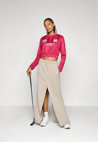 Felpa corta rosa con logo, abbinata a pantaloni beige stile avvolgente. Scarpe da ginnastica bianche e mazza da golf completano l'outfit.
