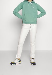 Sudadera con capucha verde menta y bolsillo frontal, combinada con jeans blancos de tiro alto y zapatillas deportivas coloridas.