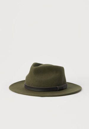 Chapeau fedora à large bord de couleur vert olive avec une bande en cuir noir autour de la base, posé sur un fond blanc.