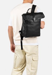 Sac à dos en cuir noir avec un design à rabat, fermeture éclair, strap de marque et bretelles ajustables. Possède une texture lisse.