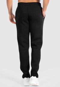 Černé tepláky s elastickým pasem, bočními kapsami a hladkým texture, spárované s bílými sportovními botami.