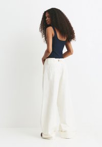 Vrouw met lang krullend haar, gekleed in een marineblauwe mouwloze top en wijde hoge witte broek, staande tegen een witte achtergrond.