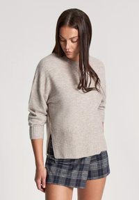 Jasnobeżowy sweter dzianinowy z ribbowanym dołem i mankietami, noszony na tle ciemnej kraciastej spódniczki mini. Sweter ma luźny fason oraz boczne rozcięcia.