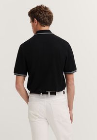 Schwarzes Polohemd mit flachem Kragen und kontrastierenden weißen Streifen an den Ärmeln, kombiniert mit weißen Hosen, zeigt die Rückansicht.