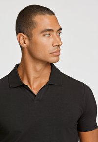Svart polo skjorta med kort krage och V-ringad design. Tyget verkar slätt med en åtsittande silhuett. Modellen har kort, mörkt hår.
