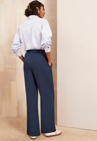 Hellblause Button-Up-Hemd mit hochgekrempelten Ärmeln, kombiniert mit dunkelblauen, weit geschnittenen Hosen mit elastischem Bund und Seitentaschen. Weiße Sneaker.
