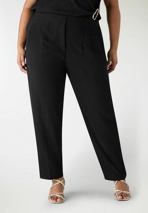 Donna con pantaloni neri a vita alta e gamba affusolata con pieghe frontali e sandali beige con tacco e cinturini, in piedi davanti a uno sfondo semplice.