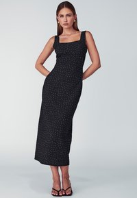Crna midi haljina s kvadratnim dekolteom i širokim naramenicama, ukrašena bijelim točkicama, od lagane tkanine, sa pripijenim siluetom.