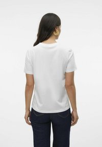 T-shirt en coton blanc à manches courtes, avec un col rond et une coupe droite, associé à un jean bleu foncé. Boucles d'oreilles en or visibles.