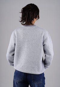 Grauer Sweatshirt aus einem weichen, strukturierten Stoff mit geripptem Ausschnitt und Bündchen, präsentiert sich in einer lockeren Silhouette mit überschnittenen Schultern. Darunter sind blaue Jeans sichtbar.