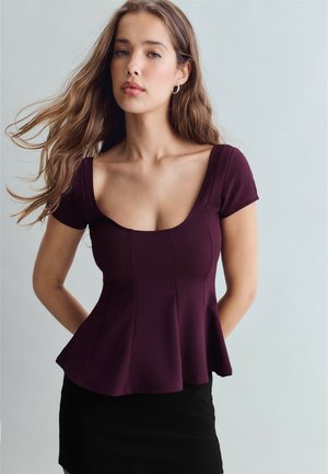 Jeune femme aux longs cheveux bruns portant un haut violet moulant à manches courtes et une jupe noire, debout devant un fond gris clair uni.