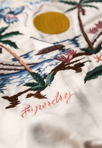 Biały materiał haftowany w palmowe drzewa, słońce, żaglówkę oraz kwiatowe wzory w odcieniach zieleni, żółci, różu i brązu. Napis "Superdry" w różowej nici.