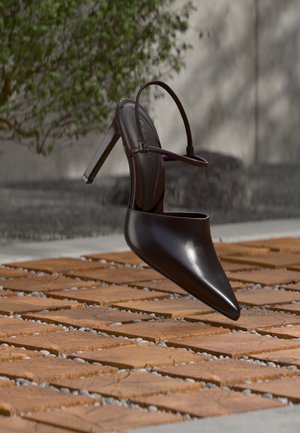 Glanzende zwarte hoge hak mule met spitse neus en enkelriem, zwevend boven een oppervlak met terracottategels en grindvoegen.