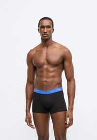 Mustad bokserpüksid sinise vöökohaga, millel on "Tommy Hilfiger" logo, valmistatud pehmest, venivast materjalist. Modell poseerib püsti.