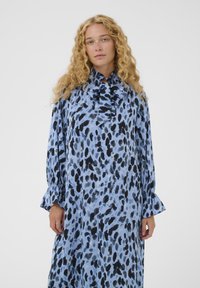 Femeie cu păr creț blond, purtând o rochie albastră cu mâneci lungi, împodobită cu pete abstracte negre și gri, și cu o cravată de guler volanată.