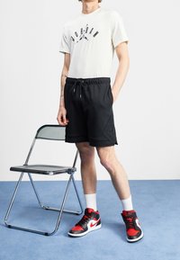 Vit t-shirt med svart "Jordan" logotyp, svarta shorts, vita strumpor, röda och svarta högstövlar, metallisk hopfällbar stol i bakgrunden.