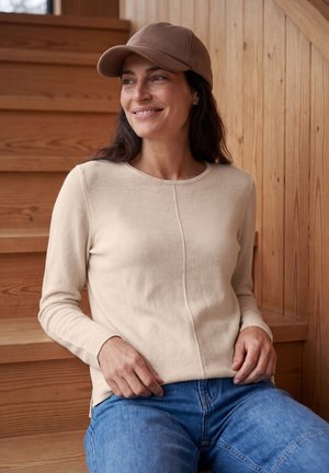 Mujer sonriendo y sentada en escaleras de madera, vistiendo un suéter beige, gorra marrón y vaqueros azules mirando hacia un lado.