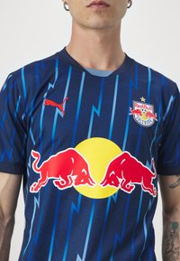 Il logo dei Red Bulls su una maglietta sportiva blu navy presenta motivi a fulmini blu e accenti rossi. Maniche corte, scollo girocollo, tessuto traspirante.