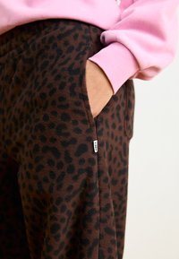 Braune Fleece-Jogginghosen mit einem schwarzen Leopardenmuster, ausgestattet mit einer Seiten Tasche und einem kleinen weißen Etikett an der Naht. Ein leichter pinker Sweaterärmel ist sichtbar.