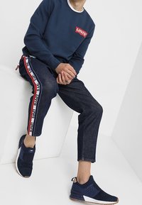 Námořnický svetr s červeným logem Levi’s, kombinovaný s tmavými džínami s bočními pruhy a námořnickými teniskami s bíl ými akcenty.