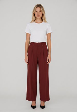 Mujer rubia de pie, con una camisa blanca de manga corta, pantalones marrones de pierna ancha y talle alto, y zapatos de tacón negros, frente a un fondo gris.