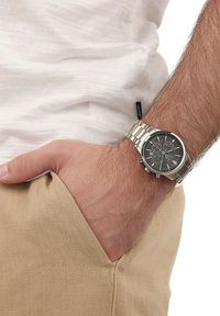 Männliche Hand mit beigem Hosentaschen, die eine silberne Metallarmbanduhr mit schwarzem Zifferblatt und Chronograph auf einem behaarten Handgelenk trägt, weißes Hemd ist sichtbar.