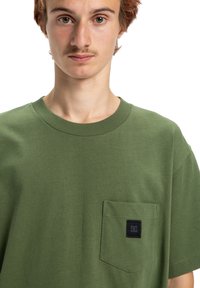 Olivově zelené bavlněné tričko s kulatým výstřihem a krátkými rukávy. Na levé straně na hrudi se nachází kapsička s černou nášivkou loga.