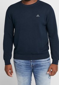 Marineblauwe gebreide trui met een gevarieerd patroon, ronde hals en GANT-logo op de borst. Strakke manchetten en tailleband. Draagt lichtgewassen jeans.